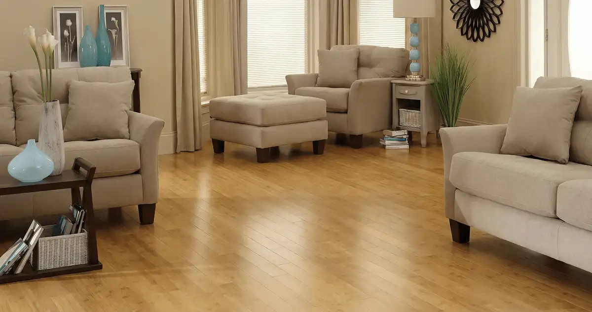 A living room with professionally installed bamboo flooring, beige sofas, a matching ottoman, a side table with a blue lamp, vases, framed pictures, and large windows with blinds and curtains in Dover, DE